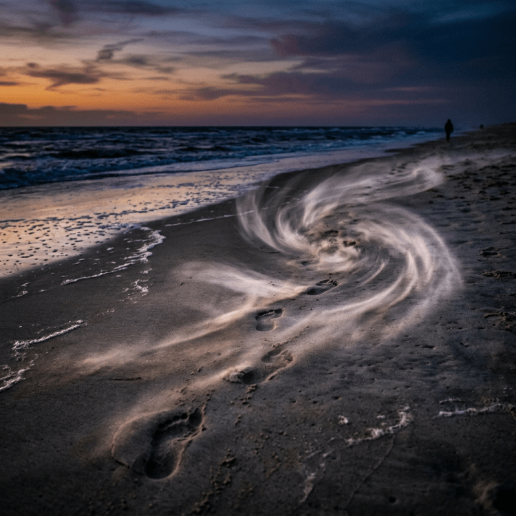 Swirling sand patterns around footprints on a beach at sunset with ocean waves